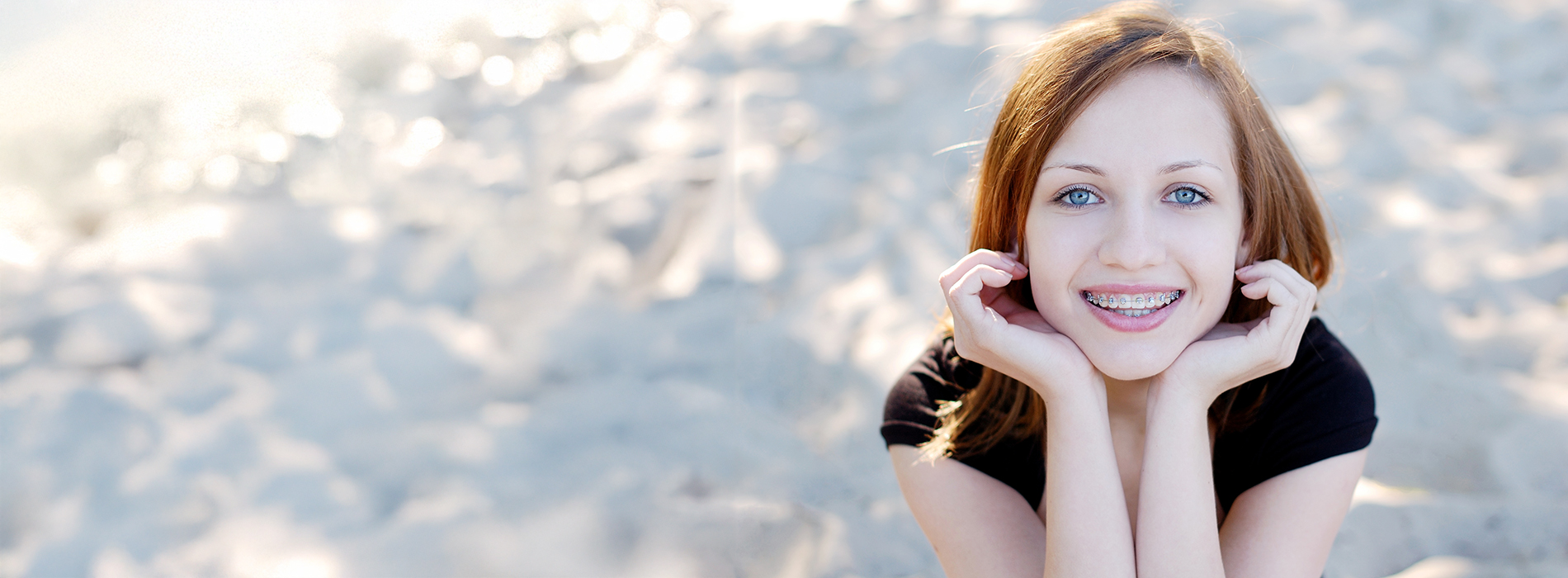 A young woman poses on a sandy beach, leaning on her hand with a smile, wearing a black top, against a backdrop of the ocean.